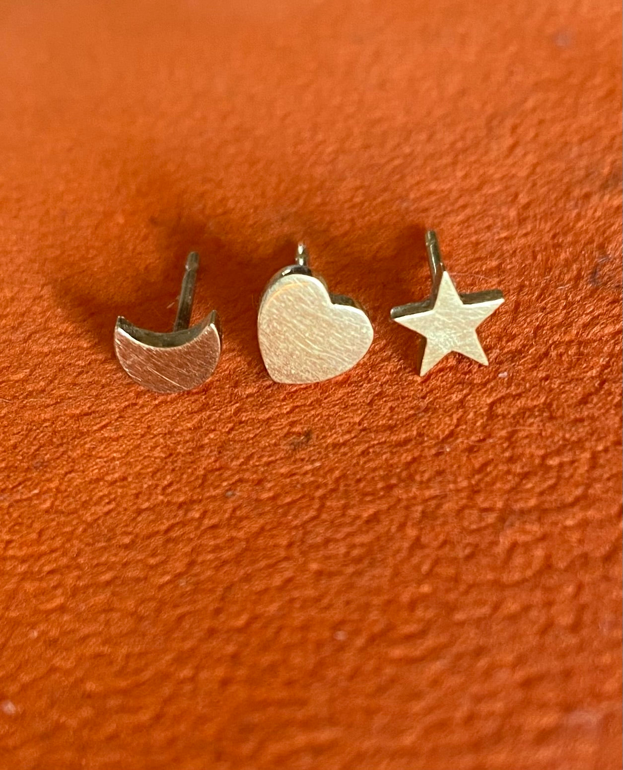 Three small metal earrings shaped like a half-circle, heart, and star on an orange surface.