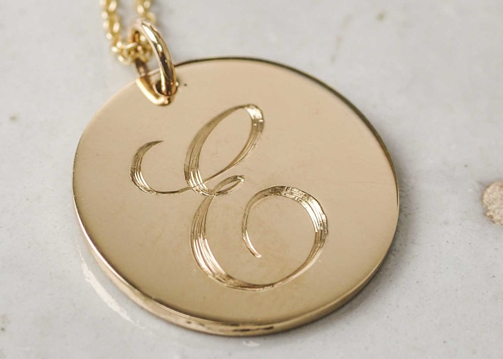 A solid gold pendant necklace with an engraved initial 'E' in script font, on a chain, displayed on a marble surface.